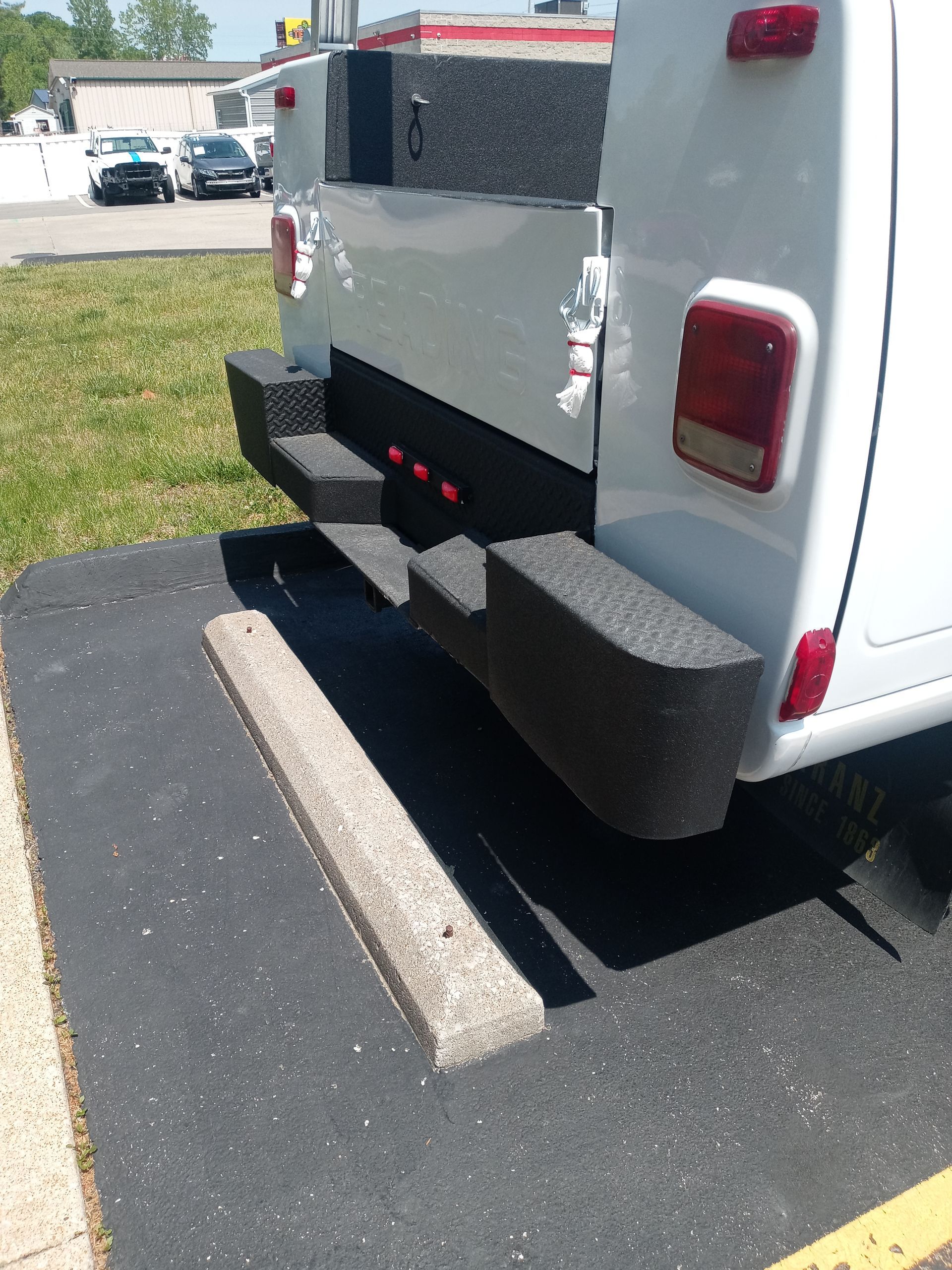 White utility vehicle rear with black bumper and step. Parked near a concrete curb.