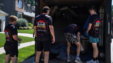 Four people in matching black shirts load items into a garage from the driveway.
