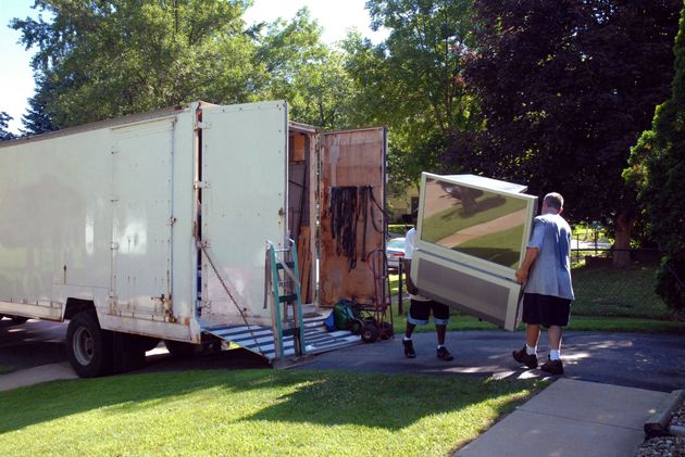Two people move a large white appliance into a parked moving truck on a sunny residential street.