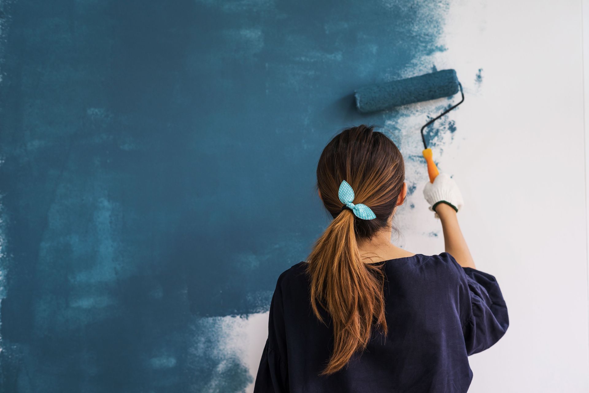 Woman painting a wall blue with a paint roller, wearing a blue shirt and gloves.
