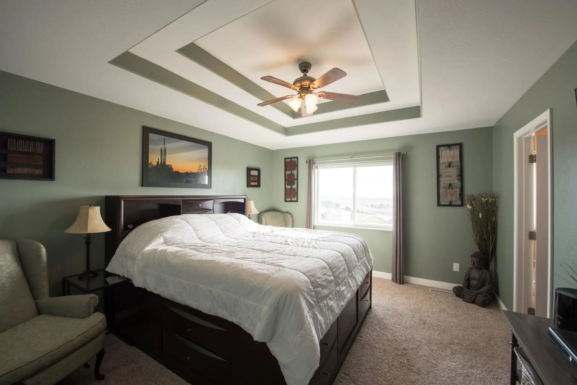 Bedroom with a large bed, two armchairs, a window, and decorative wall art. Beige walls and a tray ceiling.