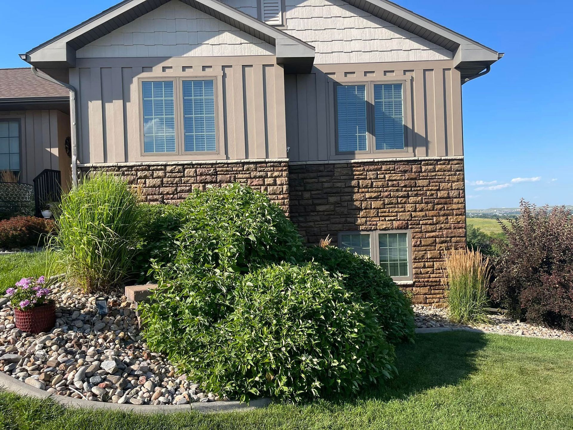 House with tan siding and stone facade, two windows, bushes in front.
