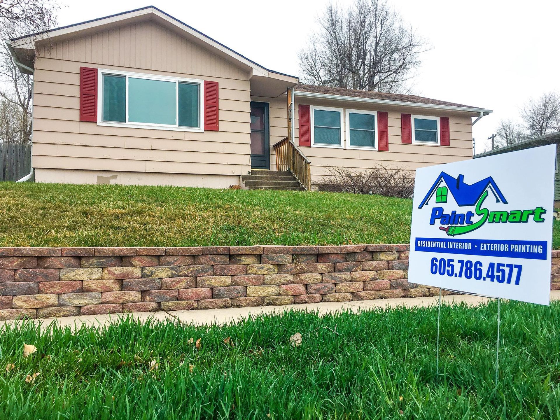 House with tan siding, red shutters, and Paint Smart sign on a grassy lawn; exterior painting services.