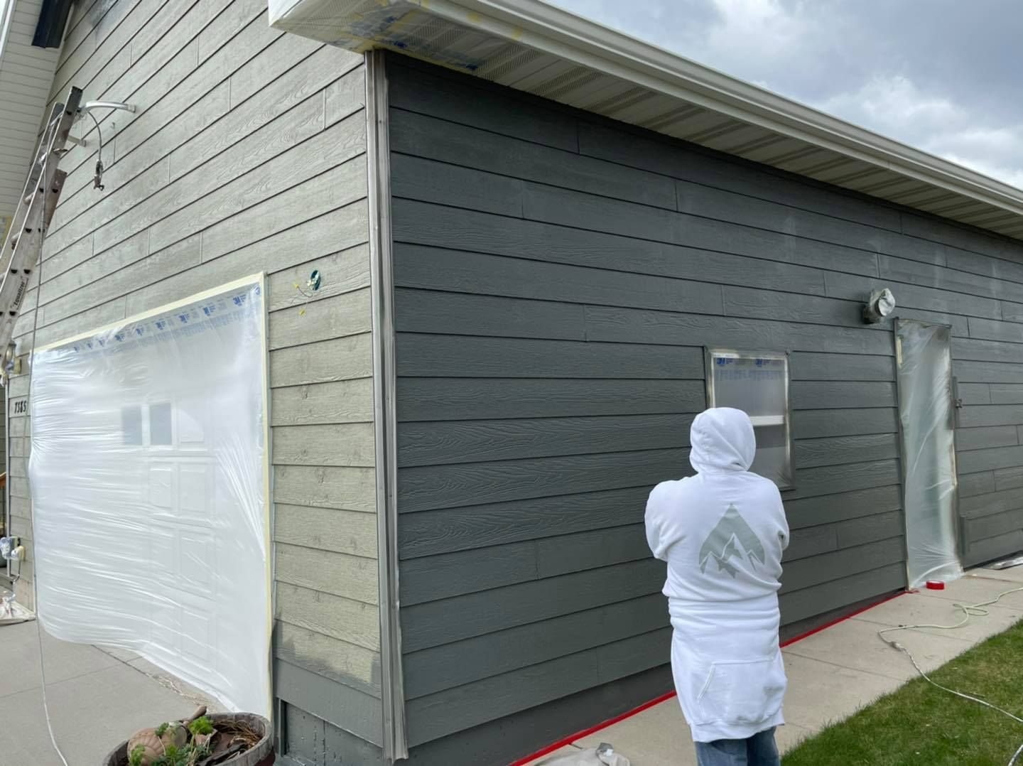 Person in protective suit painting dark gray siding on a building. White plastic covers garage door and window.