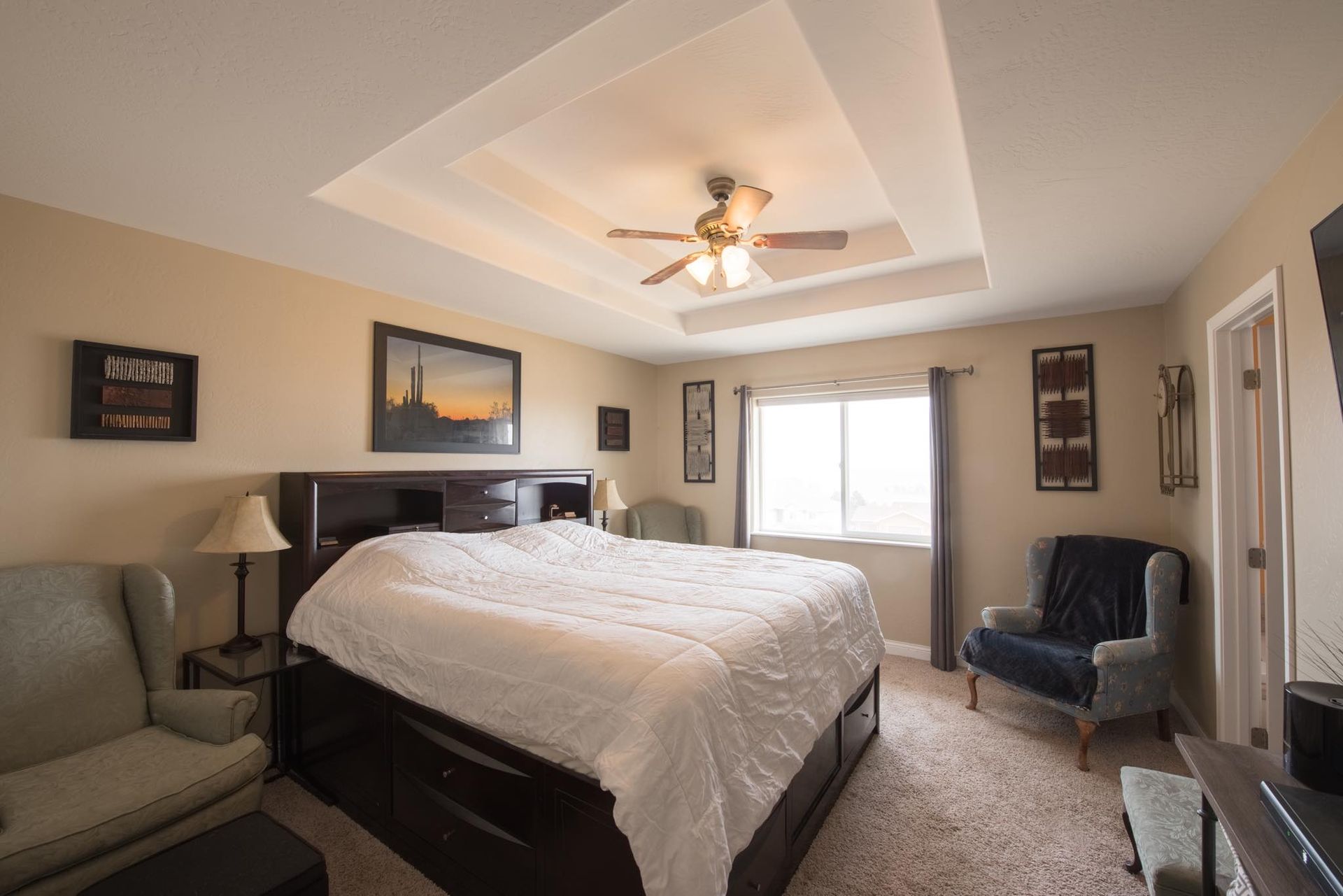 Bedroom with a large bed, two armchairs, a window, and decorative wall art. Beige walls and a tray ceiling.