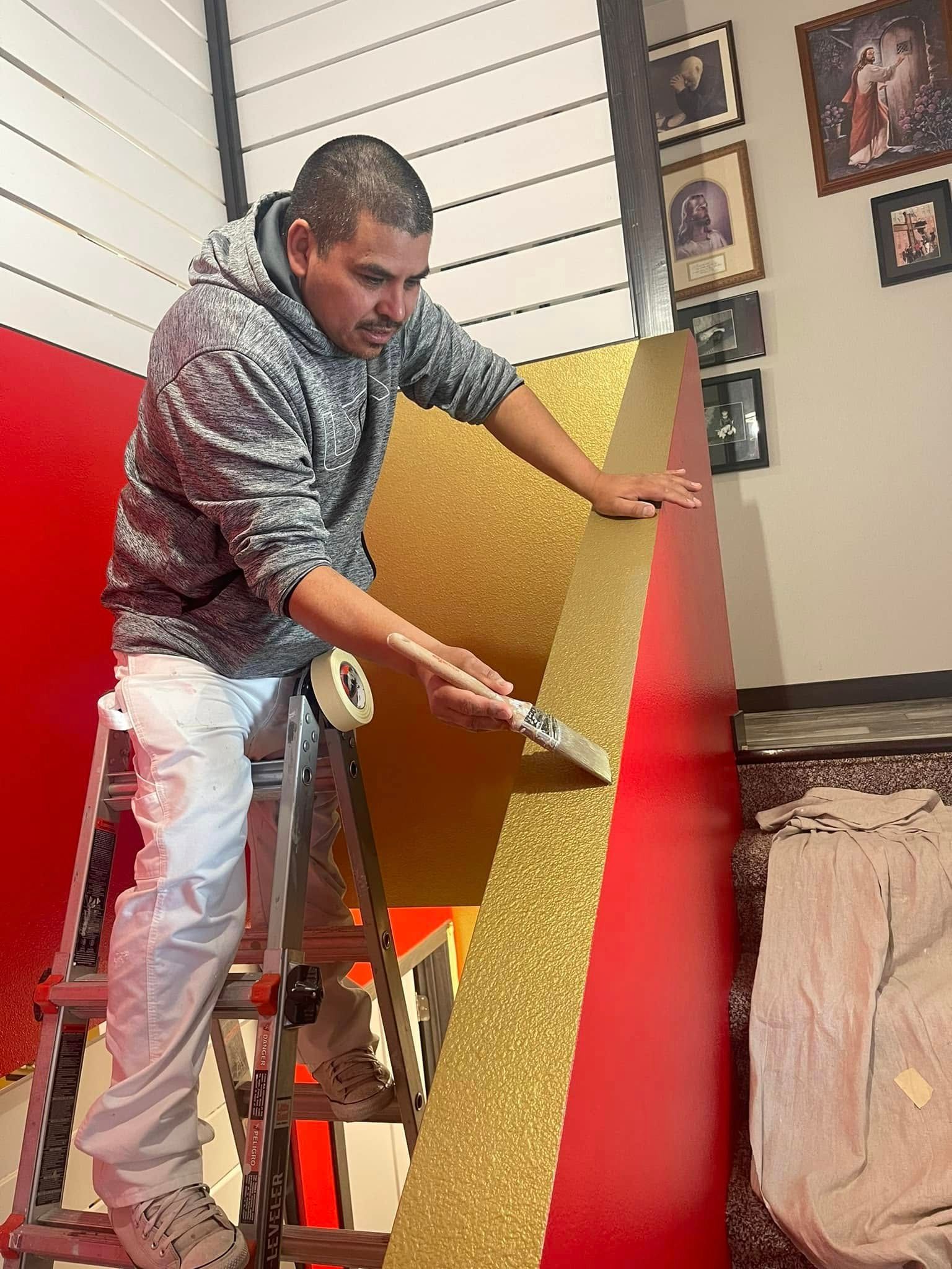 Man on a ladder applying gold glitter tape to a red surface. Indoor setting.