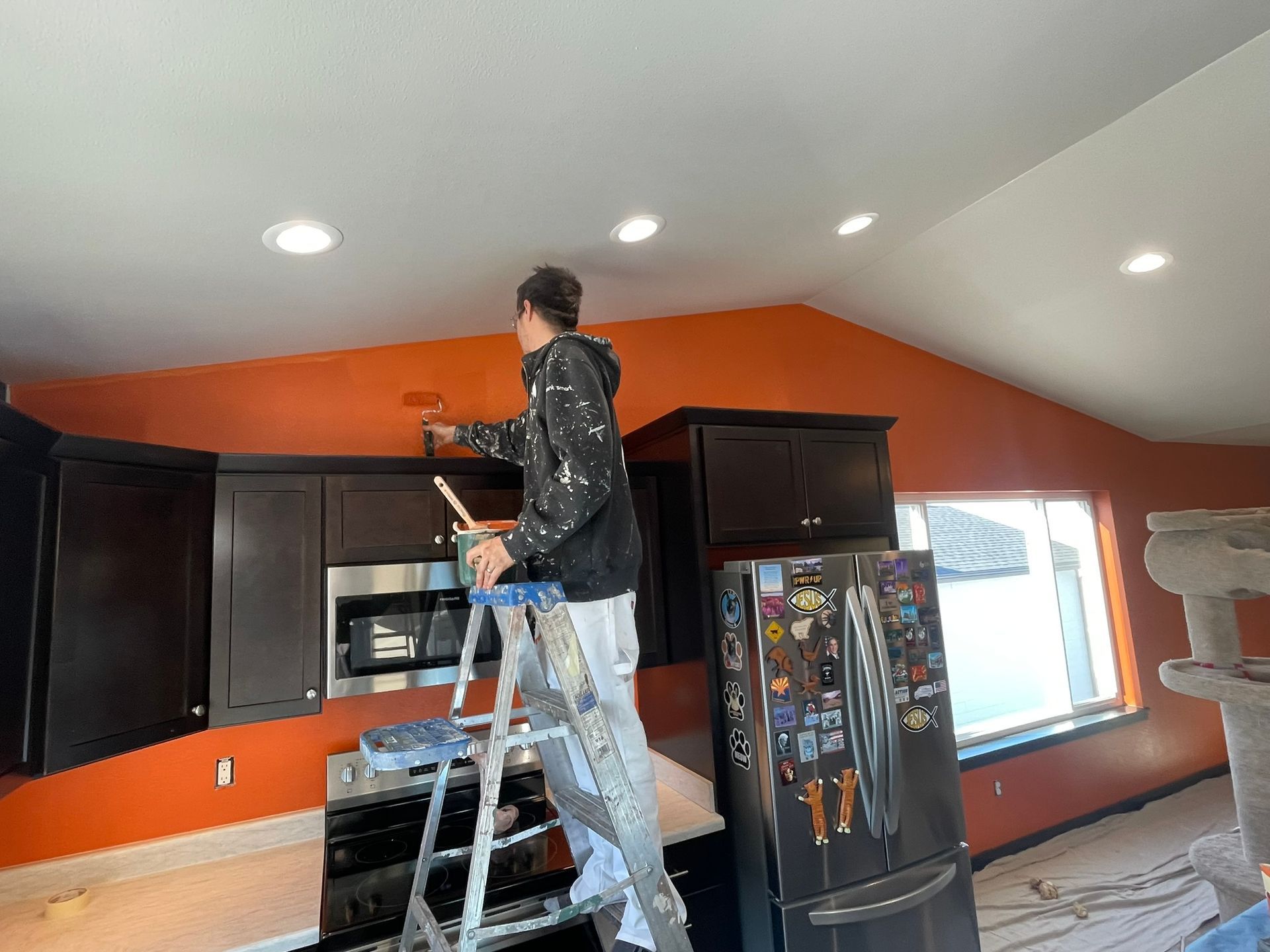 Person on a ladder painting a kitchen ceiling orange; cabinets, microwave, and refrigerator visible.
