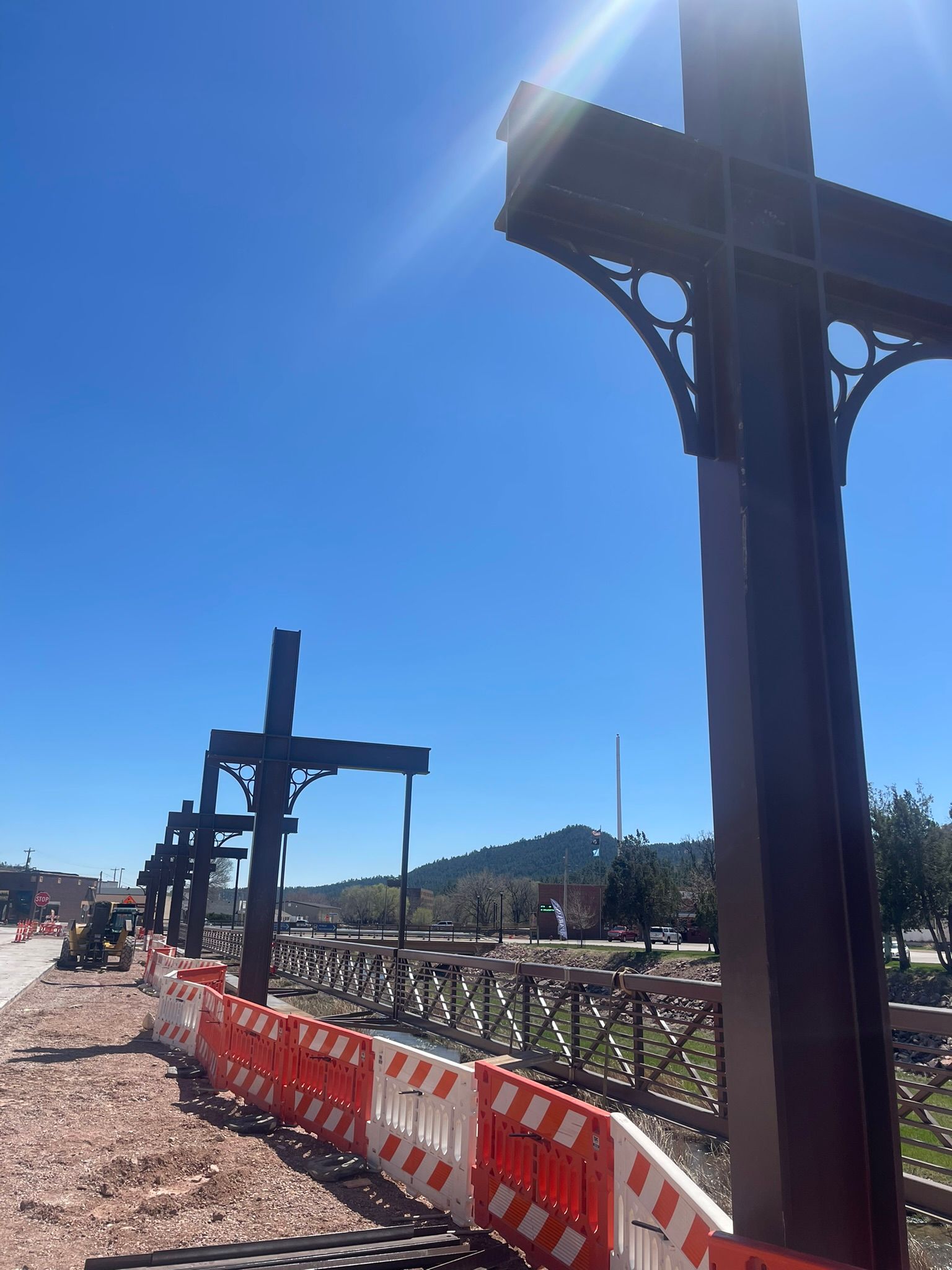Row of large metal crosses under construction, behind orange and white barriers, against a blue sky.