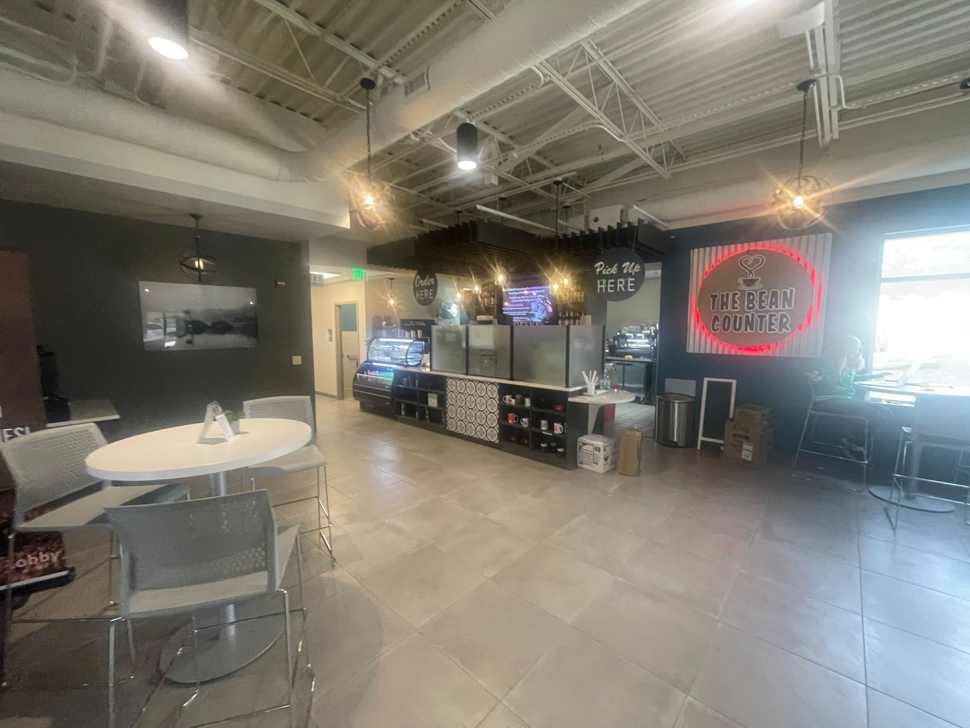 Interior of a coffee shop: tables, chairs, counter with pastries and coffee machines, neon sign.