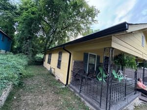 Yellow house with black gutters, front porch, and green lawn; person sits on porch.