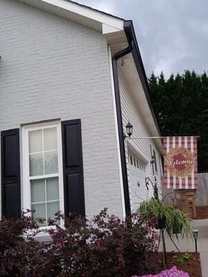 Gray brick house with black shutters and a welcome flag.