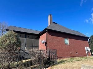Red brick house with dark roof, chimney, satellite dish, and black wrought iron fence against a clear blue sky.