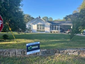A white house with a carport and a sign in front on a green lawn; blue sky.