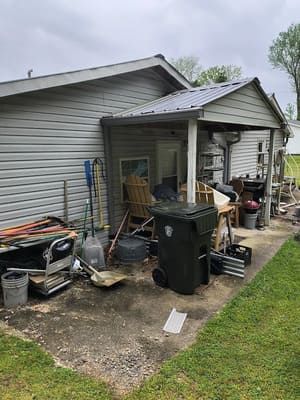 Cluttered backyard with a house, garbage bin, tools, and miscellaneous items on a concrete patio.
