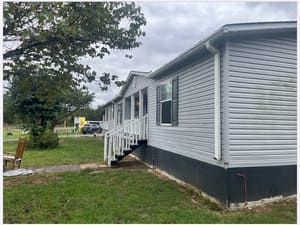 Gray-sided mobile home with black trim, steps, and a cloudy sky. Lawn and trees surround.