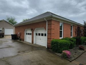 Brick garage with white doors, windows, and green bushes. Overcast sky.