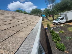 Roof with brown shingles and a white gutter next to a truck in a sunny yard.