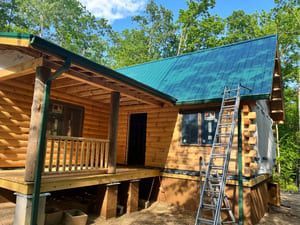 Log cabin under construction with teal metal roof and wooden porch. A-frame ladder leans against the building.