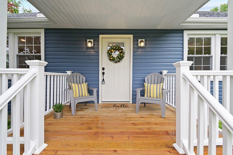 The front porch of a blue house with two chairs and a wreath on the door.