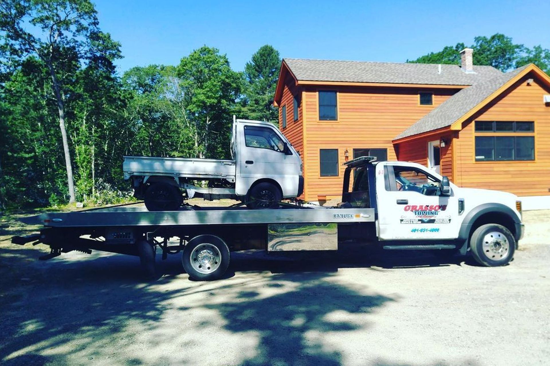 Tow truck carrying a small white truck on a flatbed, parked near a wooden house and trees.