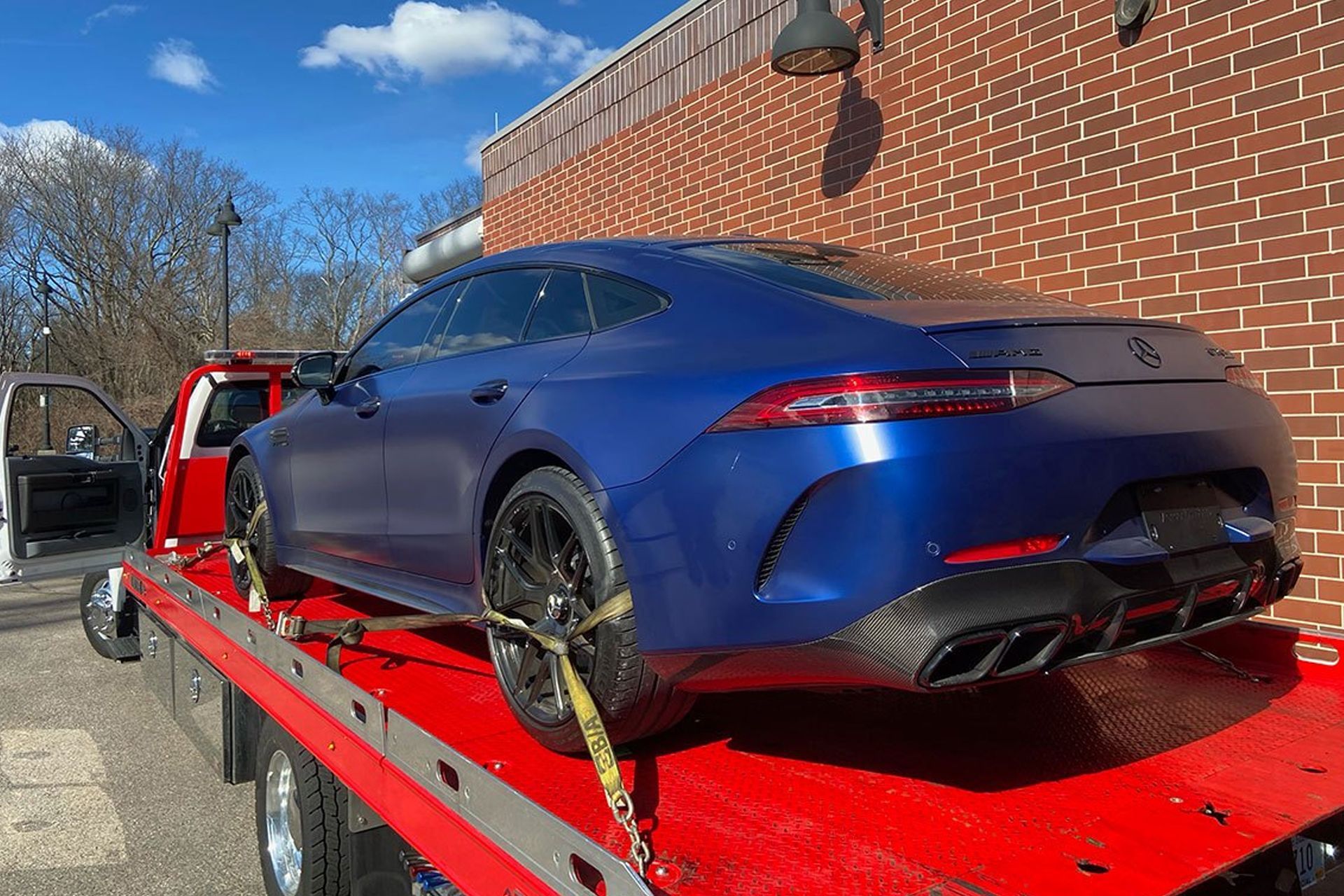 Blue Mercedes sports car on a tow truck, parked in front of a brick building on a sunny day.