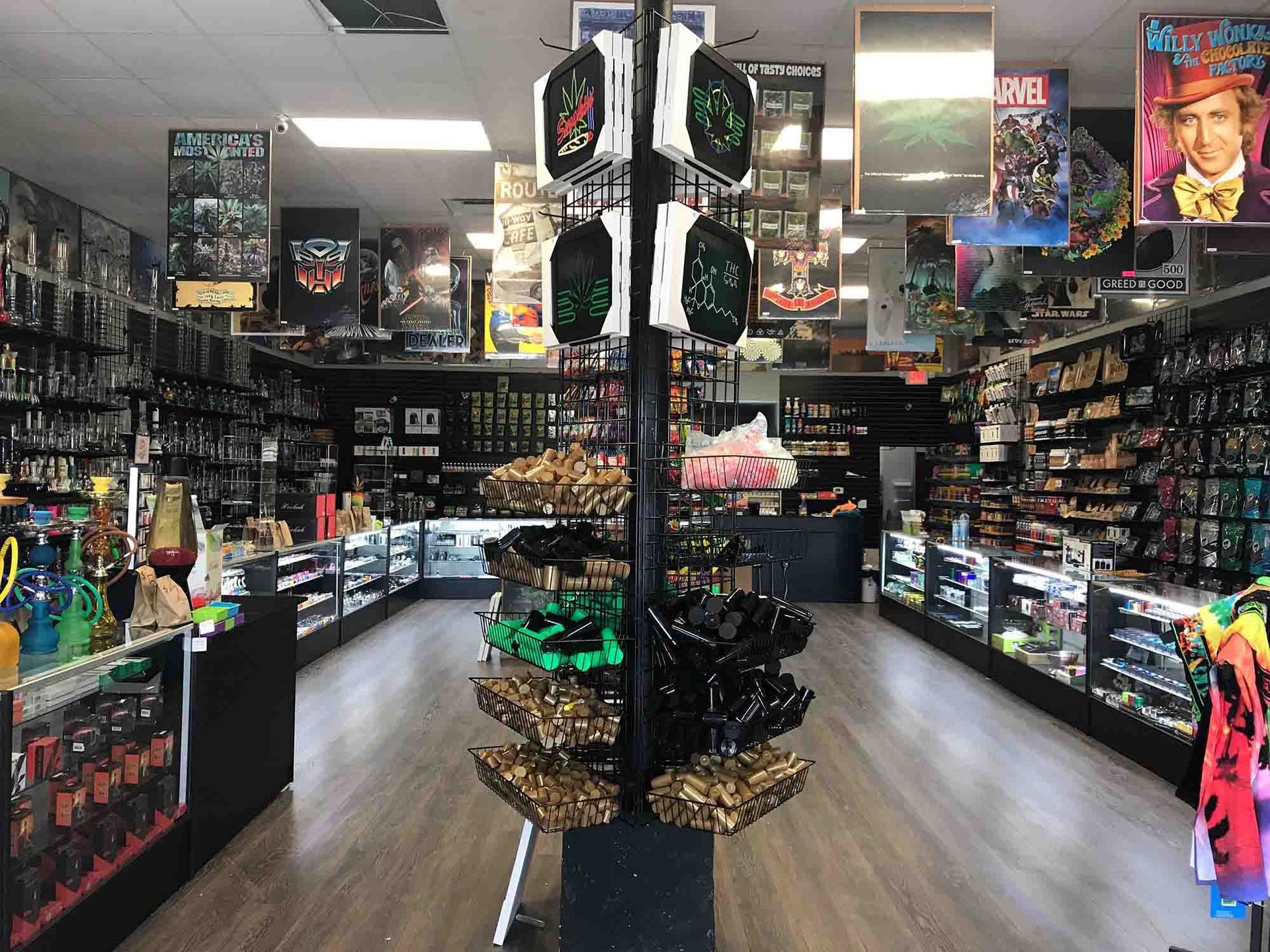 Inside a shop, items are on display. Posters hang. A display rack holds small items, with shelves.