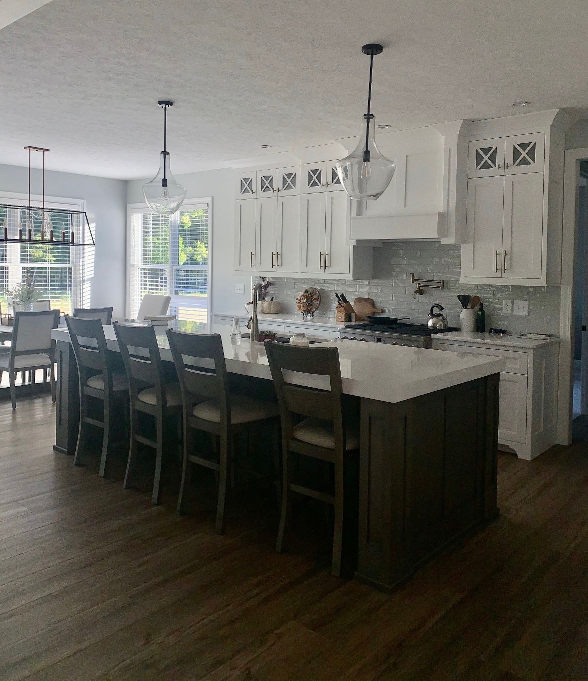A bright kitchen with a dark-wood island, white cabinetry, a marble backsplash, and four wooden stools on wood flooring.