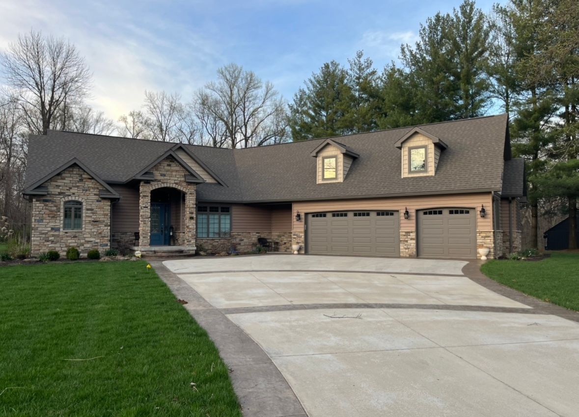 A single-story stone and siding house with a three-car garage and a wide concrete driveway in a wooded suburban setting.