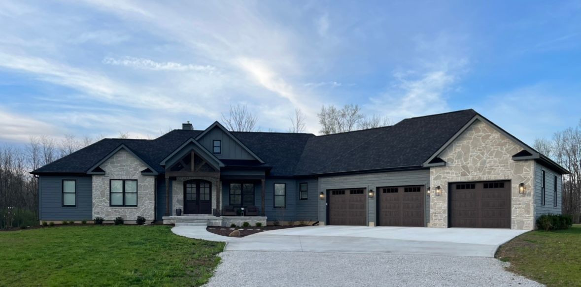 A single-story, dark gray siding and stone house with a three-car garage, large windows, and a paved driveway.