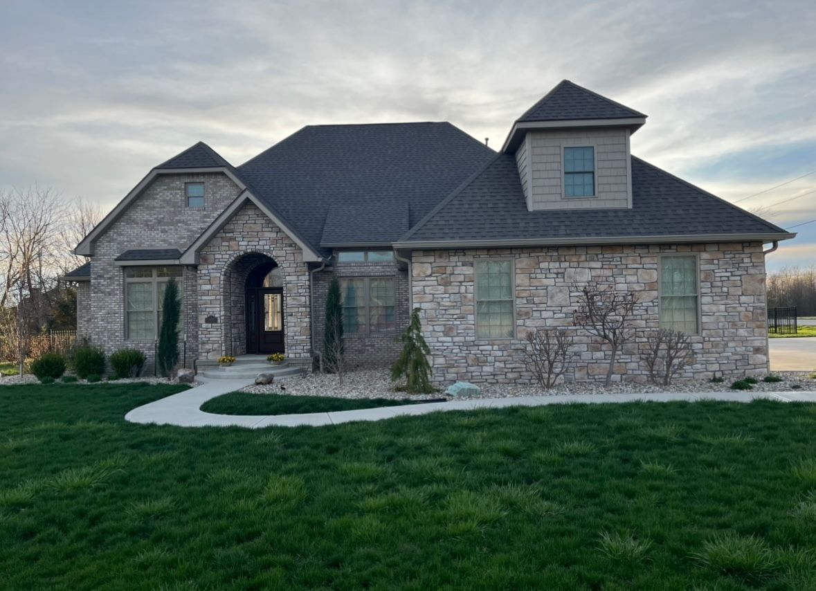 A single-story, gray stone house with a dark roof and dormer window, set behind a green lawn and concrete walkway.