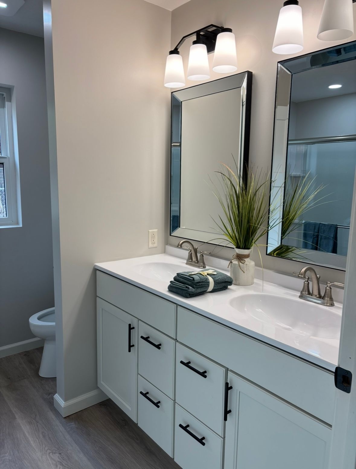 Modern bathroom with double vanity, two framed mirrors, contemporary lighting, white cabinets, and wood-look flooring.