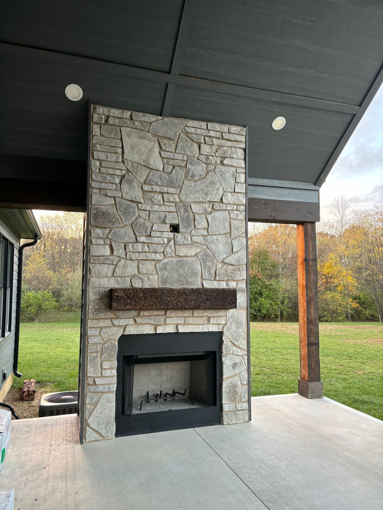 A stone outdoor fireplace with a dark wooden mantel and black firebox, set under a covered patio facing a grassy yard.