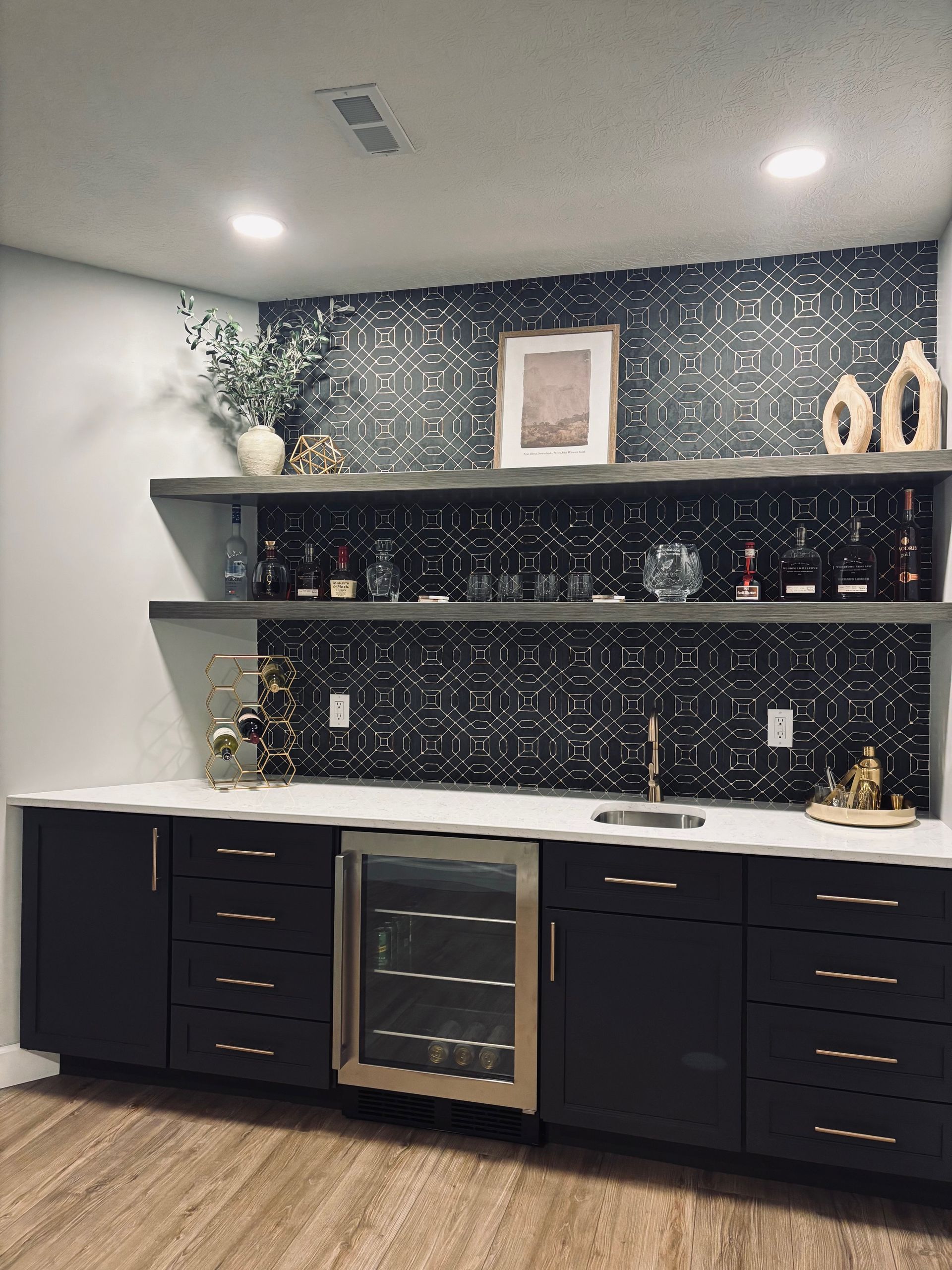 A modern wet bar featuring black cabinets, a wine fridge, white countertops, two floating shelves, and dark patterned tile.