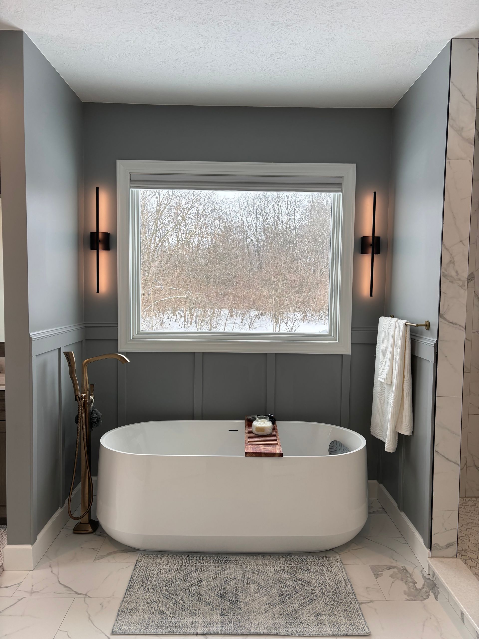 A modern bathroom with a white freestanding tub, grey board-and-batten walls, two sconces, and a window overlooking woods.