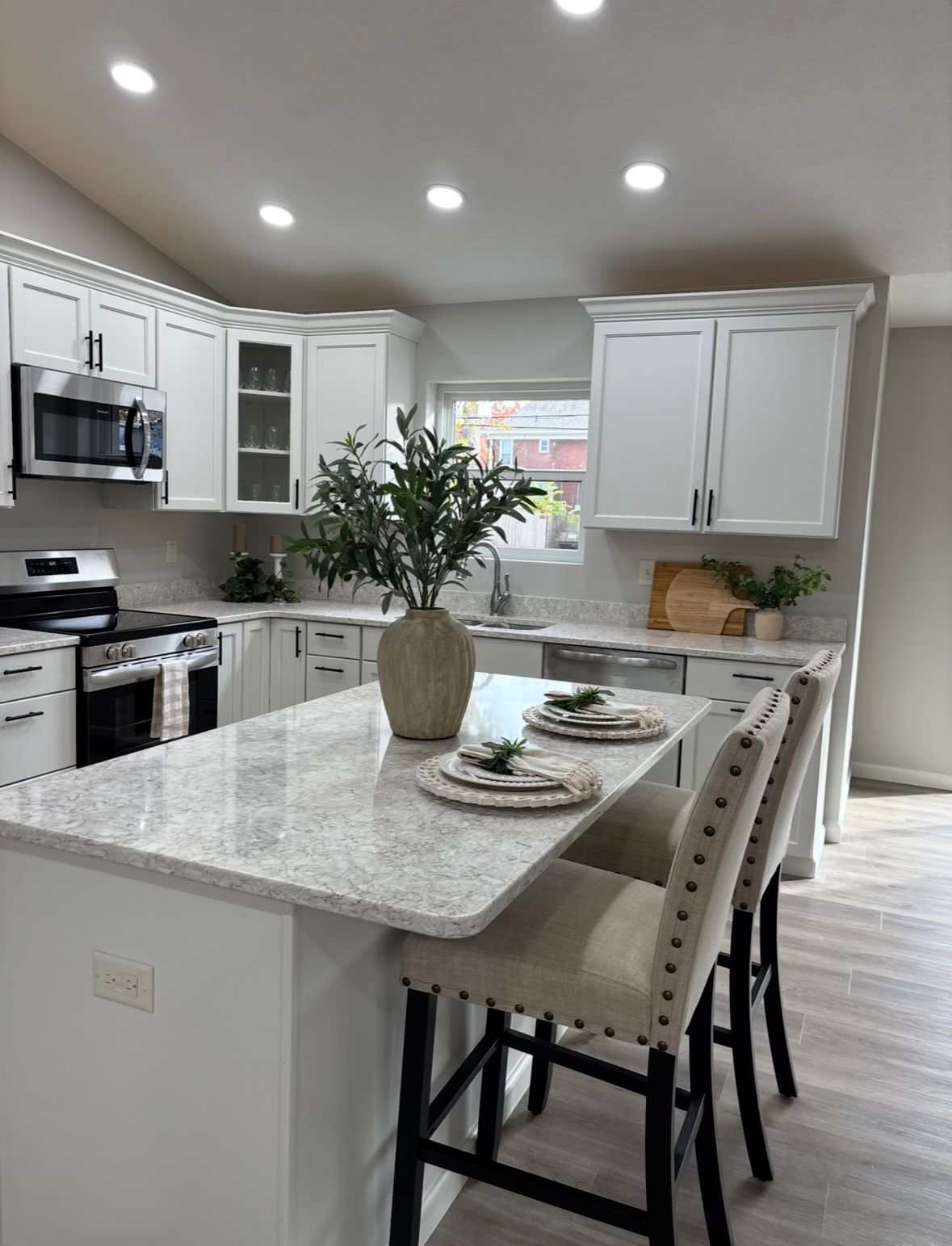 A modern kitchen with white cabinets, light-colored granite countertops, a central island, and two upholstered bar stools.