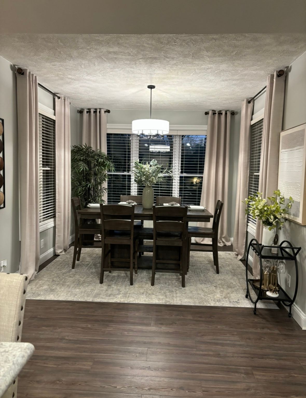 A dining room featuring a wooden table and chairs, a light gray rug, neutral curtains, and a chandelier in a bay window.