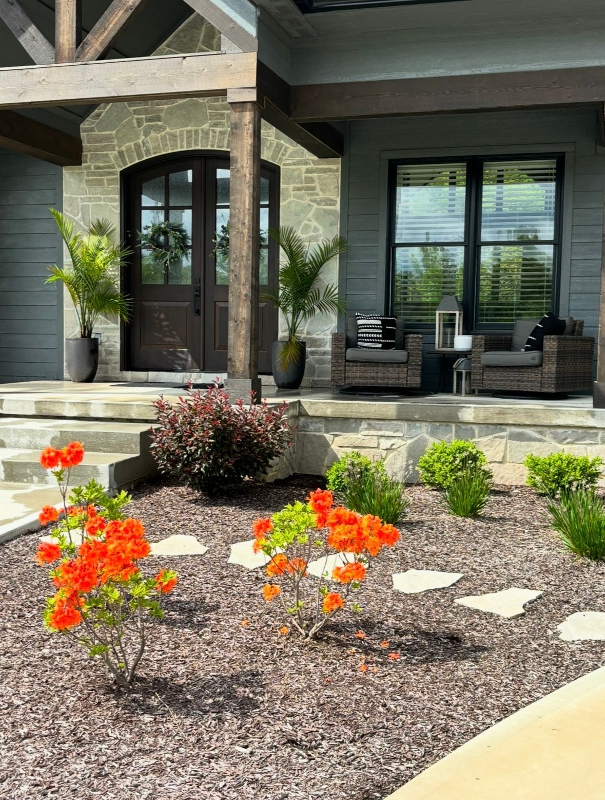 A front porch with stone accents, dark doors, and outdoor seating, featuring a garden with orange flowers and gravel.
