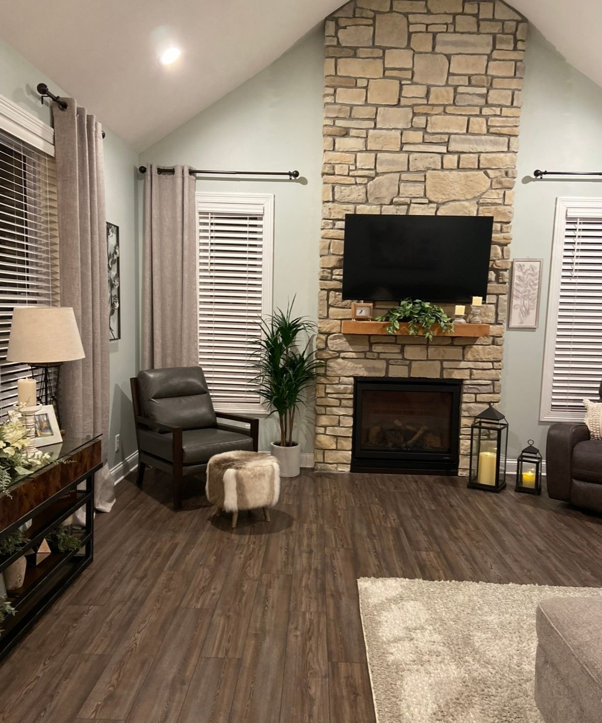 A living room features a stone fireplace with a mounted television, flanked by windows with gray curtains and blinds.