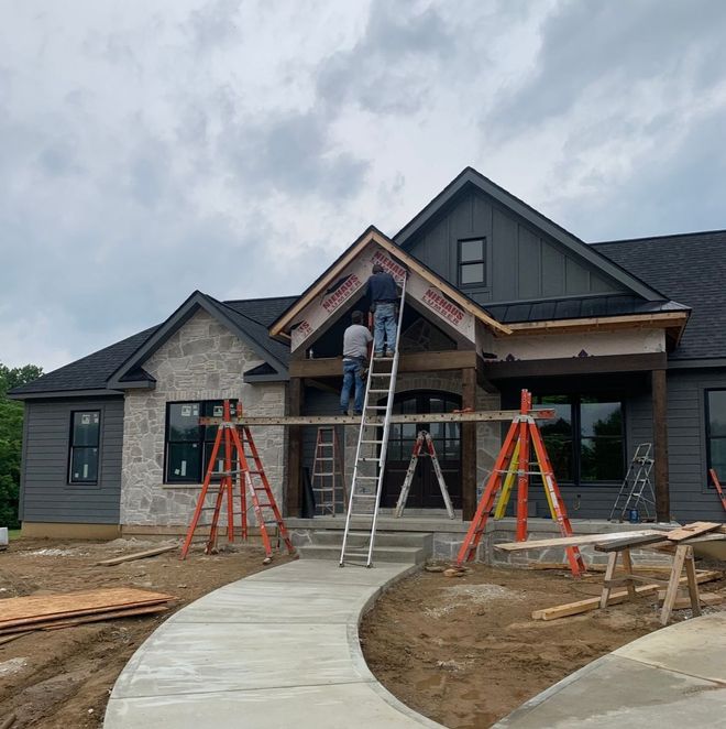 Two workers on ladders install trim on the front gable of a new house with stone and dark siding under a cloudy sky.