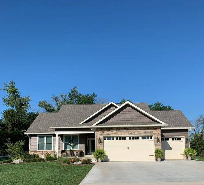 A single-story suburban house with stone siding, a gray shingled roof, a three-car garage, and a lush green lawn.