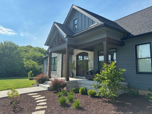 A modern farmhouse with grey horizontal siding, stone accents, a dark wood-pillared porch, and a stone walkway.