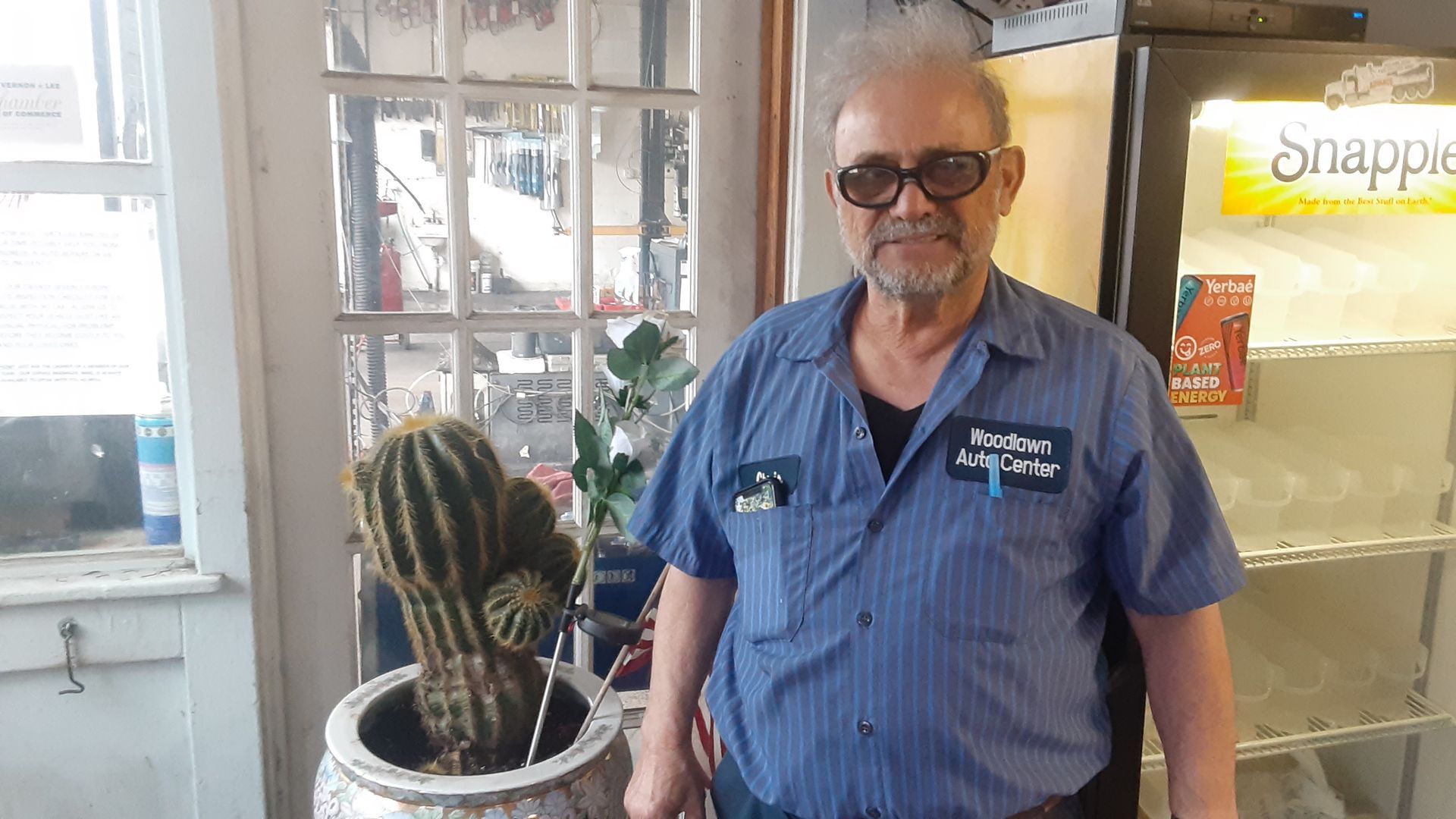 Man in blue shirt and sunglasses standing indoors beside a cactus plant near a glass door