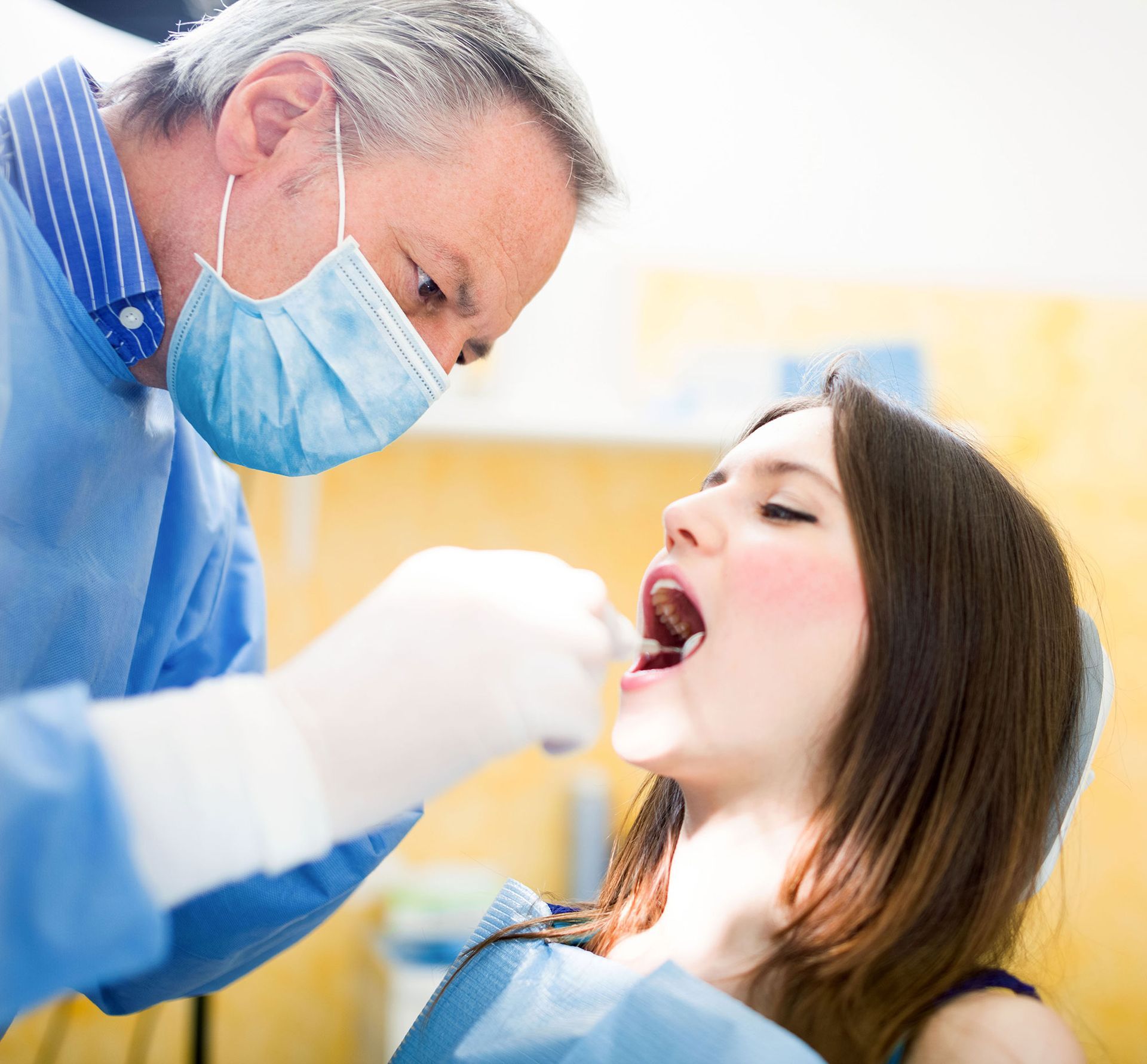 Dentist examining patient's teeth with tools in a dental office. The patient has open mouth, and the dentist is wearing a mask.