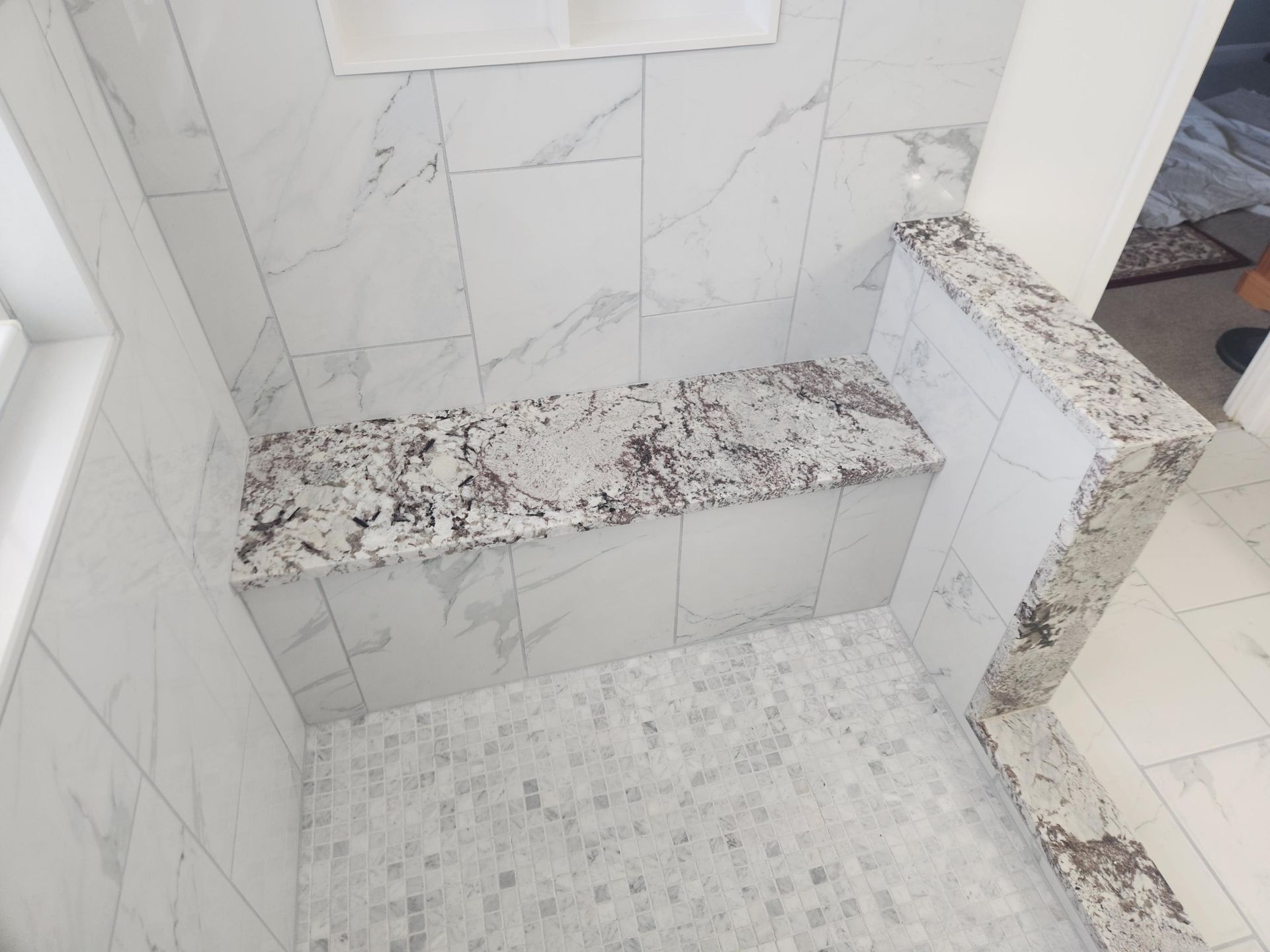 A shower with white marble-look wall tiles, a granite bench seat and top ledge, and small light gray mosaic floor tiles.