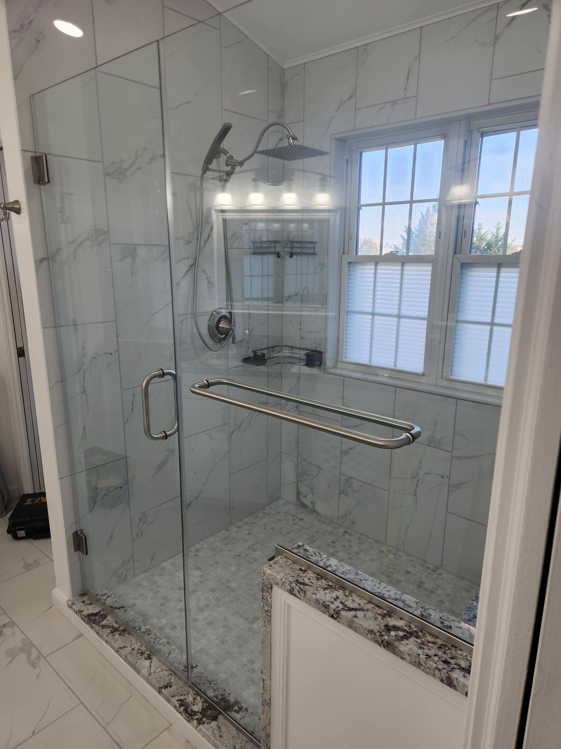 A walk-in shower with marble-look tiles, a glass door, a rain showerhead, and a granite-topped seat by a grid-pane window.