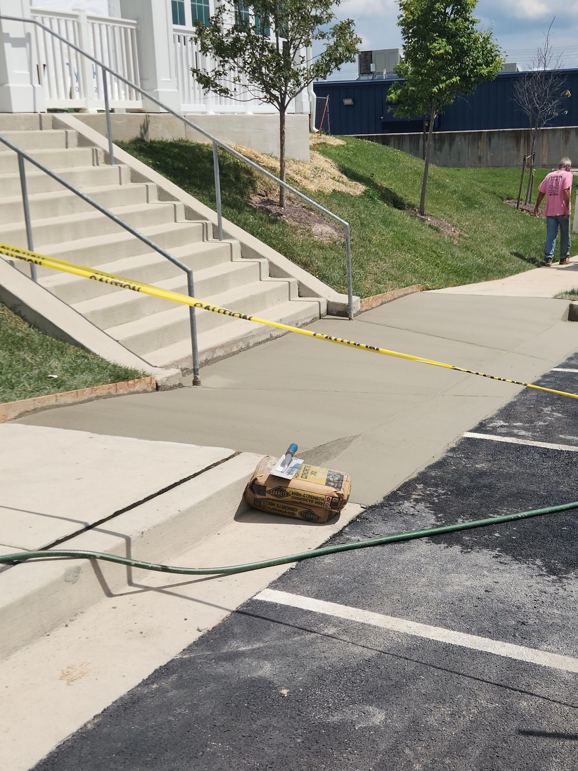 A freshly poured concrete sidewalk leads to building steps, blocked off by yellow caution tape with a person nearby.