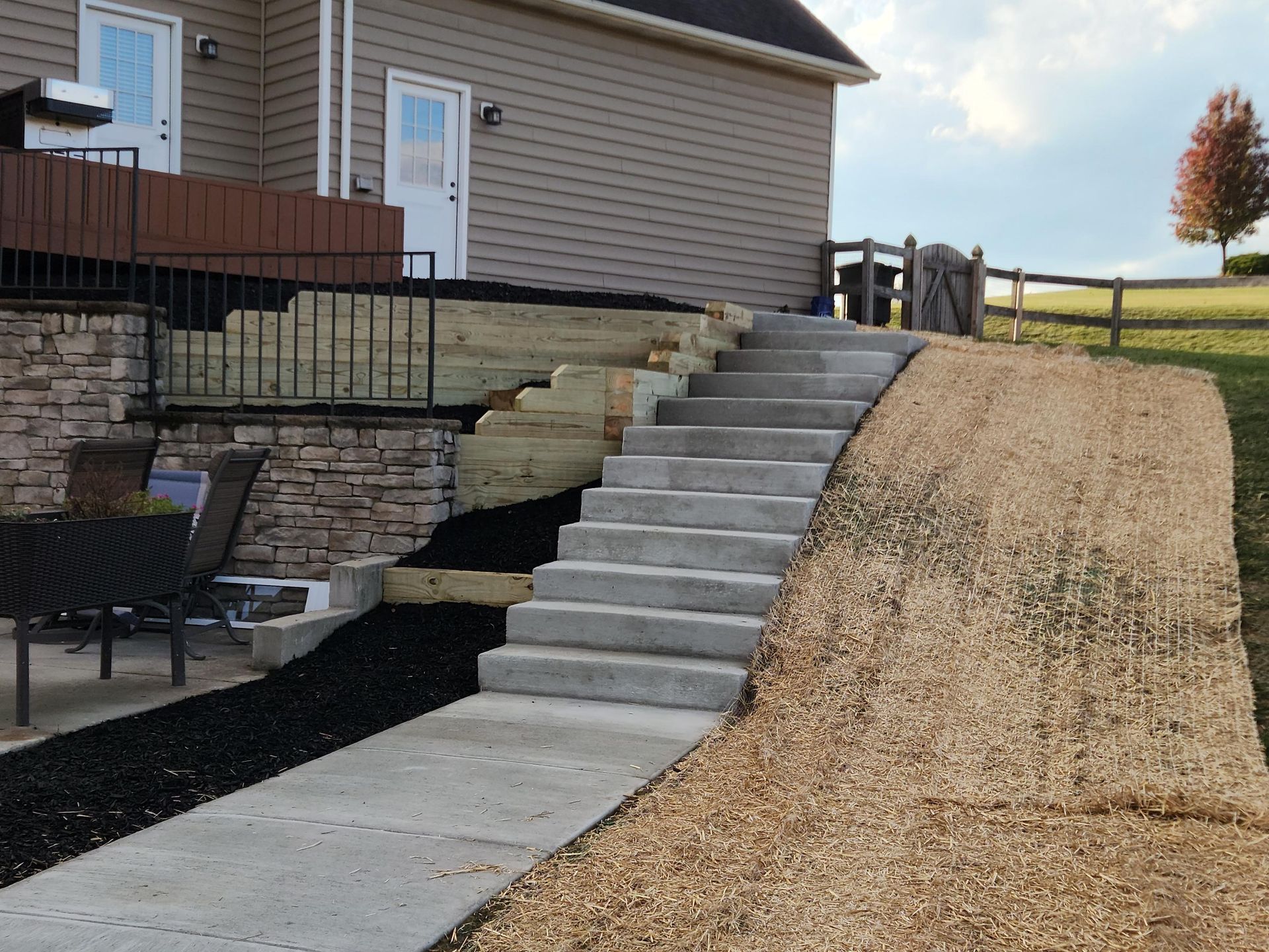 Concrete stairs lead up from a patio beside a house with a retaining wall and a newly seeded slope on the right.