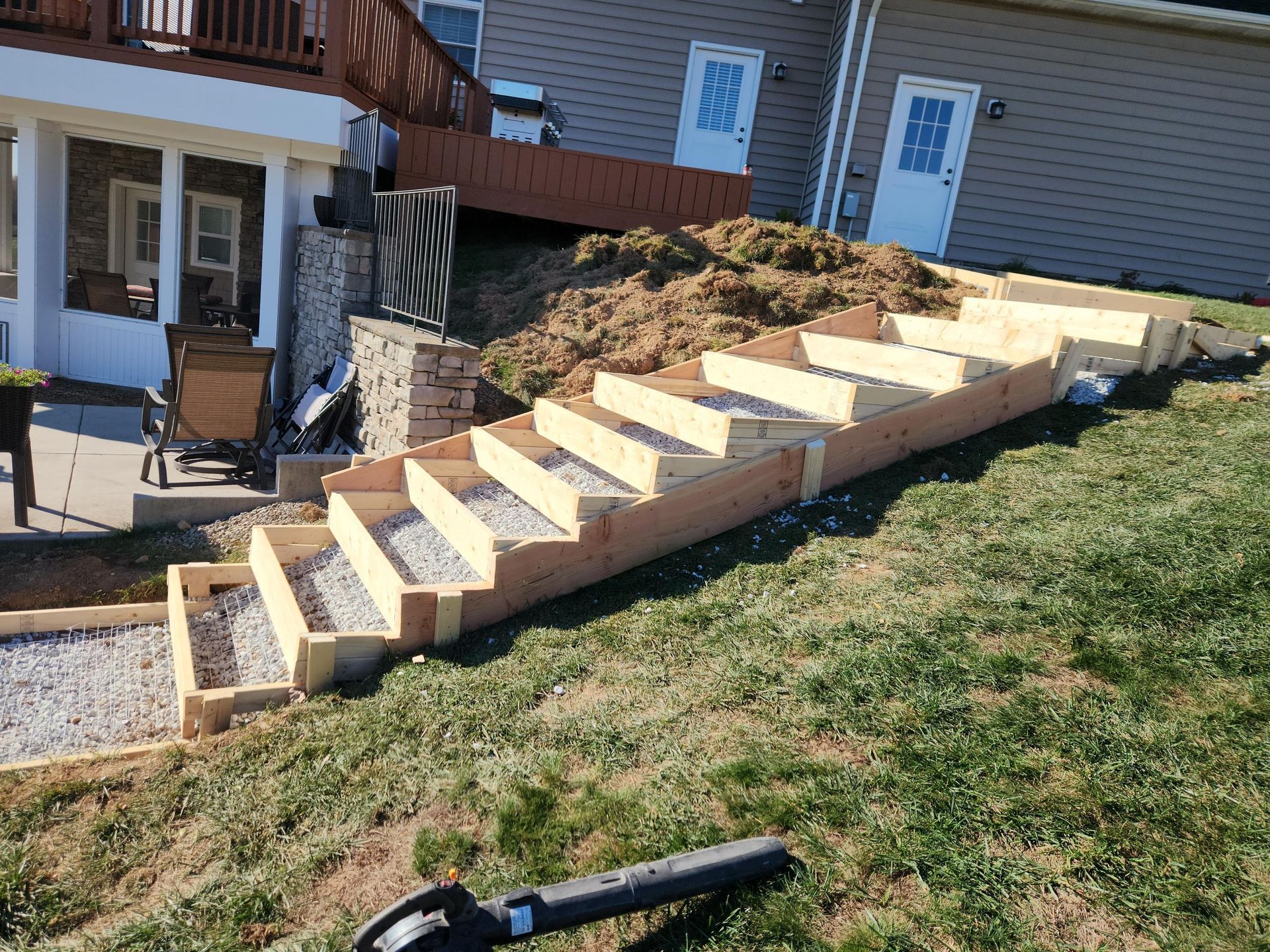 Wooden outdoor steps under construction on a grassy hill, leading up to a house with a deck.
