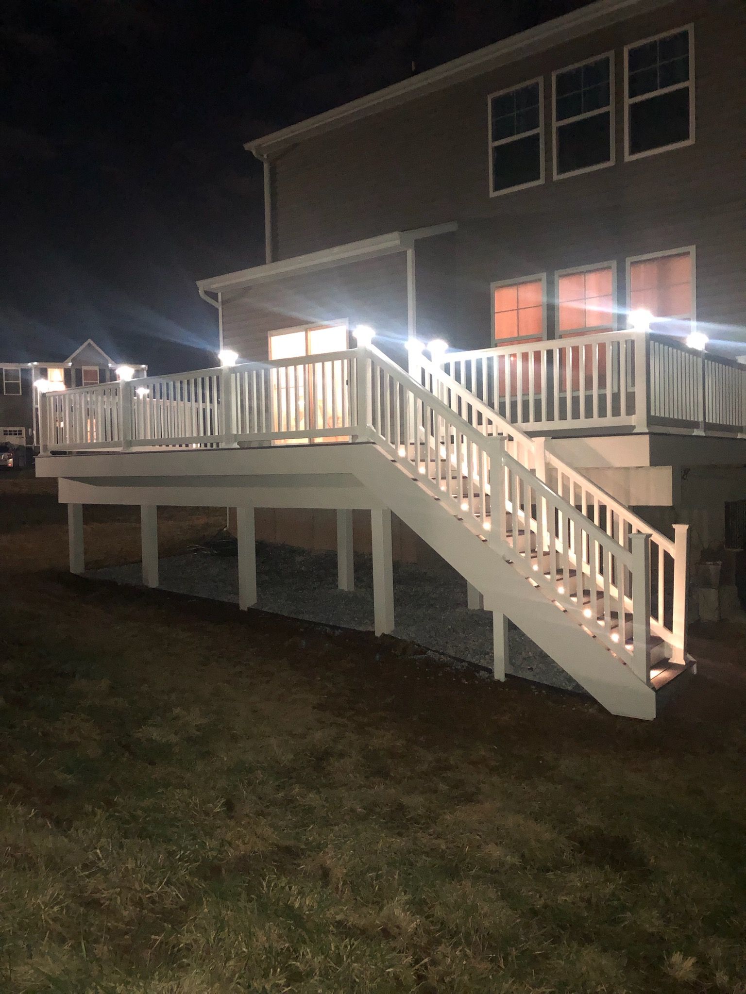 A wooden deck with white railings and illuminated post cap lights at night, attached to the back of a house.