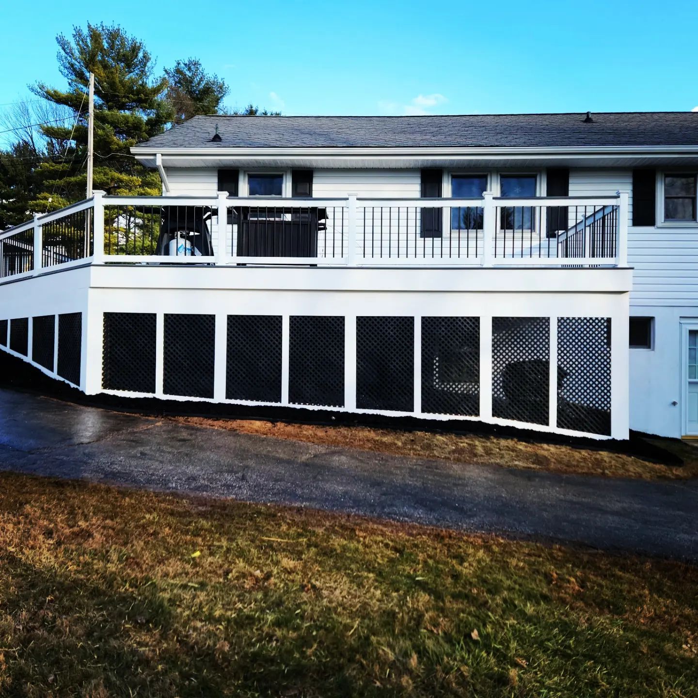 A white house with a large, elevated deck featuring black lattice skirting and black railings, set above a paved driveway.